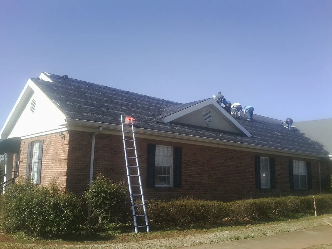 Crew tearing shingles off a brick ranch roof with a ladder set — Metal Roof Repair work by our crew in New Ross