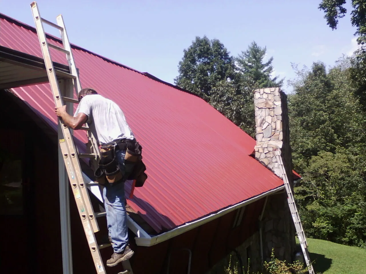 Roofer on a ladder beside a red metal roof and stone chimney — Soffit & Fascia Repair by Roofing Brothers in New Ross